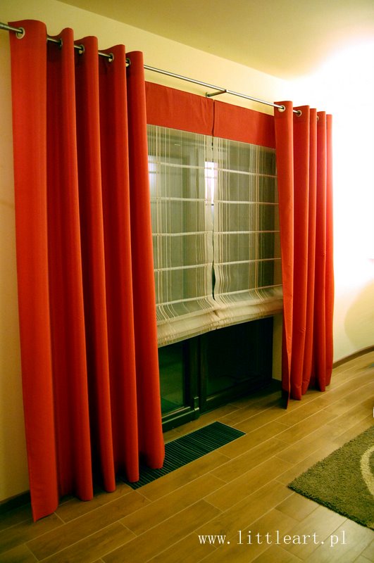 Czerwone zasłony na metalowym karniszu z przelotkami, okno z roletą rzymską w tle, drewniana podłoga i zielony dywanik w rogu, zdjęcie z lekkim prześwietleniem.